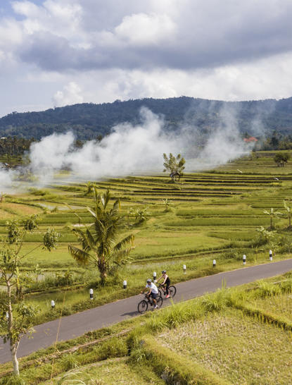 Cyclist riding along a rural road through verdant rice paddies at Amankila, with volcanic peaks and mist in the distance.