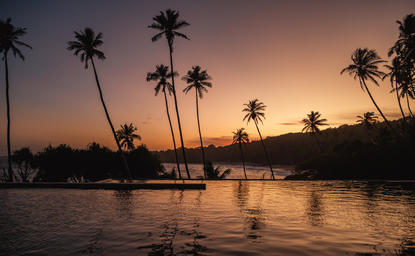 Amanwella's exterior swimming pool at sunset, with palm trees silhouetted against an amber sky and still water reflecting the light.