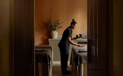 Homem em spa de bem-estar do Aman New York, apoiado em mesa de tratamento em sala serena com tons quentes.