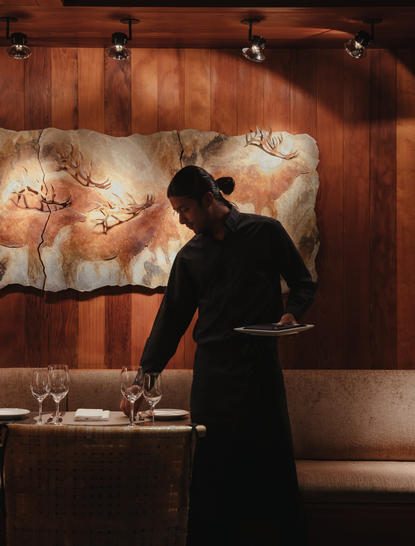 Server in black uniform arranging table settings in Amangani's dining venue, warm wood interior visible behind.