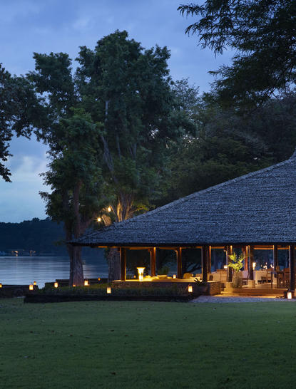 Moyo restaurant at Amanwana illuminated at dusk, overlooking the water with tropical gardens beyond.