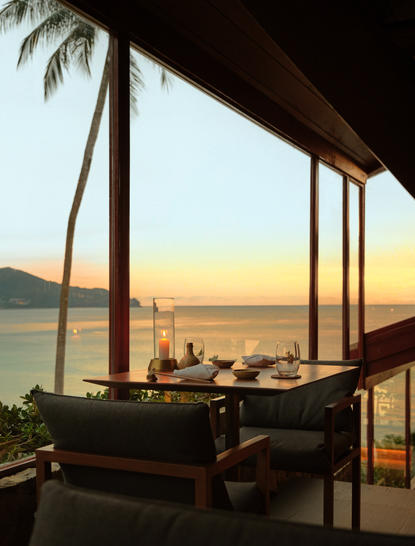 Amanpuri dining pavilion at sunset overlooking Phuket Bay, with palm fronds and calm waters visible through open windows.