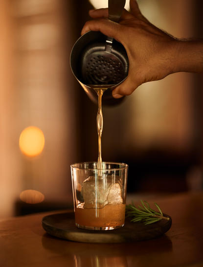Cocktail being poured at Amanjena's bar, amber liquid streaming into a glass with ice and garnish.