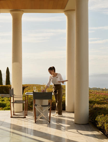 Gość siedzi przy stole w głównej restauracji Amanzoe, patrząc na krajobraz wzgórz otoczonych cyprysami.