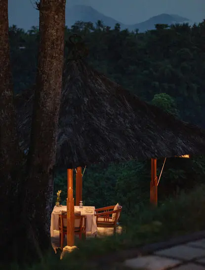 Dining bale at Amandari overlooking the Ubud valley at dusk, with wooden chairs illuminated against the darkening landscape.