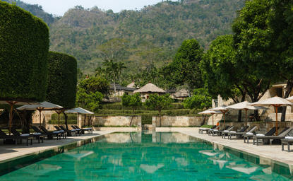 Amanjiwo's main swimming pool with emerald water, surrounded by sun loungers and cream umbrellas, set against forested hillside.