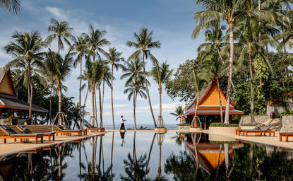 Amanpuri's wooden pavilions reflect in still water surrounded by palm trees at dawn.