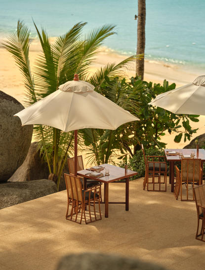 Dining terrace at Amanpuri resort overlooking the Andaman Sea, with parasol-shaded table and wooden chairs on sandy beach.
