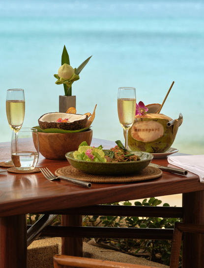 Dining table set up on a beach terrace at Amanpuri resort with champagne flutes, golden serving vessels and fresh salad, overlooking the sea.