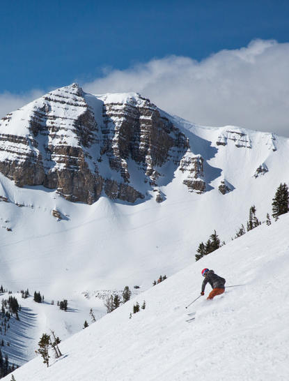 Skiers and snowboarders descend snowy slopes with snow-covered mountains and a lodge visible in the distance at Amangani.