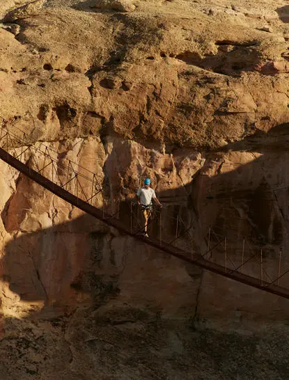 A solitary figure walks across a narrow wooden bridge suspended above a deep canyon at Amangiri, framed by towering rust-coloured rock formations.