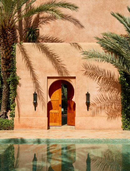 アマンジェナ・リゾートの外観。テラコッタ色の壁にヤシの木が映り、黄金色の扉と静かな水路がある。