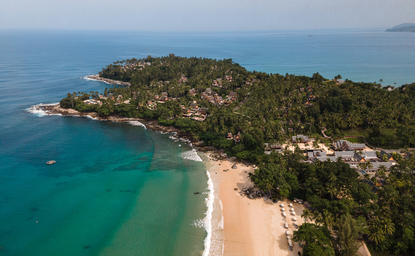 Aerial view of Amanpuri's coastal setting with sandy beach, turquoise waters, and forested headland in Thailand.