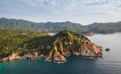 Amanoi's dramatic rocky headland rises from turquoise waters, framed by forested mountains and misty horizon.