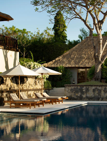 Resort swimming pool at Aman Villas at Nusa Dua, Indonesia, with thatched structures and tropical greenery.
