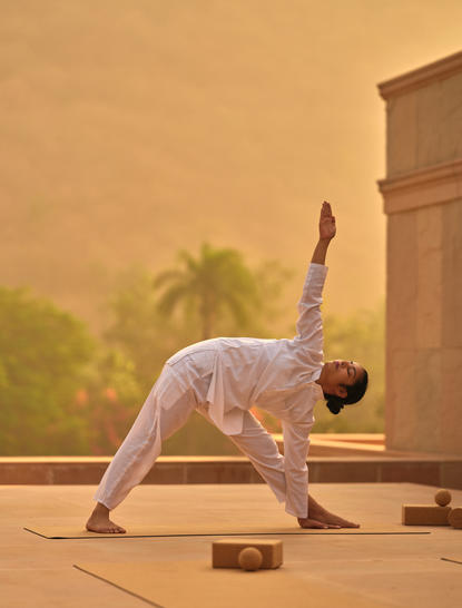 Yoga practitioner in downward-facing dog pose at Amanbagh, with warm golden light and palm trees visible beyond the terrace.