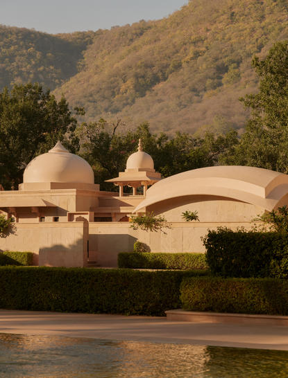 Amanbagh's accommodation pool pavilion exterior, with curved sandstone architecture beside water and forested hills beyond.