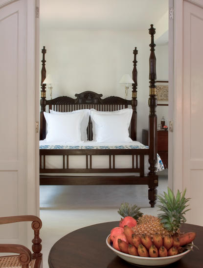 Four-poster bed with dark wooden frame in a bright bedroom at Amangalla, with fresh fruit on a table in the foreground.