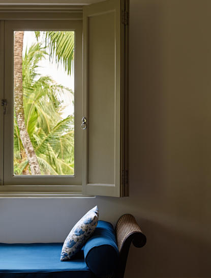 Person in blue shorts sitting on bed by window with tropical garden view at Amangalla.