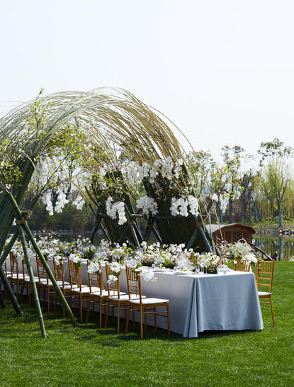 Wedding ceremony setup at Amanyangyun with curved wooden structure, blue-draped table, and white chairs on lawn.
