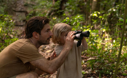 Man and child birdwatching together at Amanera, Dominican Republic, peering through binoculars at forest canopy.