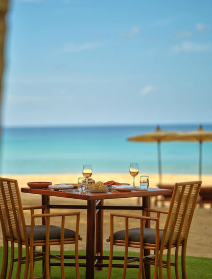 アマンプリ リゾートのヌラ ダイニングベニューで、ビーチを見渡すテーブルセットアップ。木製の椅子とテーブルが芝生に配置され、背景には紺碧の海と木製のデッキが広がる。