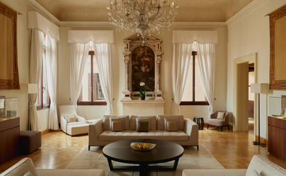 Aman Venice's Coccinas apartment living area with symmetrical seating, chandelier, and tall windows overlooking the canal.