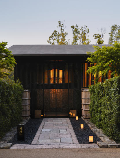 Aman Kyoto's dark timber entrance pavilion with illuminated doors, framed by green hedging and stone pathway at dusk.