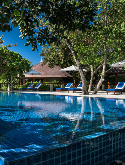 Amanpulo resort swimming pool with thatched-roof structures and mature trees reflected in calm blue water.