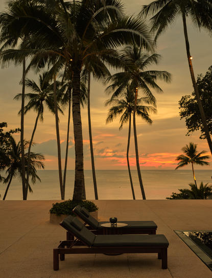Lounger on the terrace at Amanpuri resort in Thailand, overlooking the main pool at sunset with palm trees silhouetted against golden sky.
