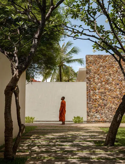 Monk in saffron robes walks through shaded courtyard at Amansara, framed by trees and stone walls.
