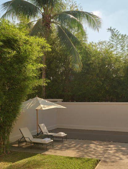 Sunlit terrace at Amantaka with loungers beside tropical greenery and palm trees.