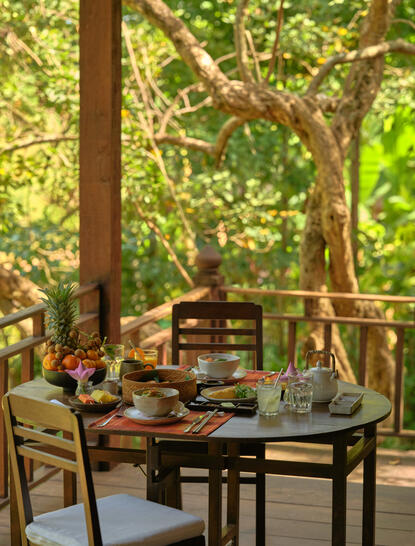 Breakfast table set on a wooden terrace at Amansara, surrounded by lush greenery and natural light filtering through the trees.