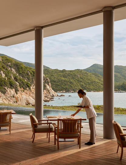 Amanoi beach club restaurant terrace overlooking coastal waters and forested hillside, with wooden dining furniture and open views.
