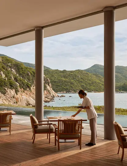Amanoi beach club restaurant terrace overlooking coastal waters and forested hillside, with wooden dining furniture and open views.