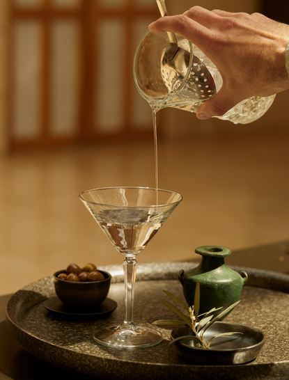 Cocktail being poured at Amanzoe resort bar in Greece, with a martini glass and bottles arranged on a dark surface.