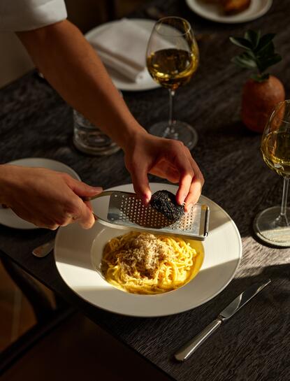 Hands plating tagliolini pasta at Aman New York's dining venue, with wine and candlelight creating an intimate atmosphere.