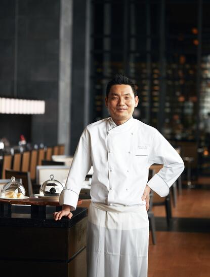 Chef Hiraki stands in Aman Tokyo's dining venue, wearing white chef's attire against an evening cityscape backdrop.
