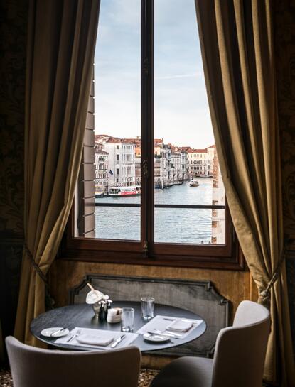 Dining table set for two at Aman Venice, framed by gold curtains with canal views through tall windows.