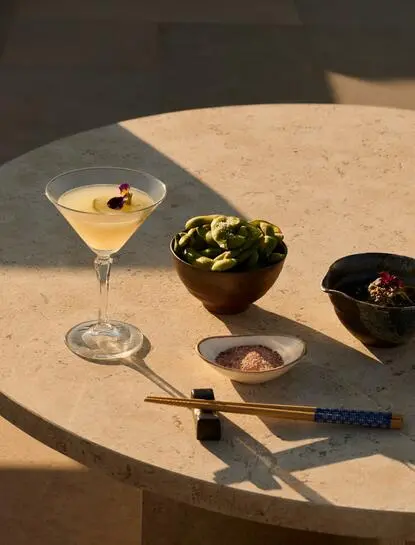 Cocktail and dining spread at Amanzoe resort, featuring a martini, fresh greens, and gold cutlery on a stone table.
