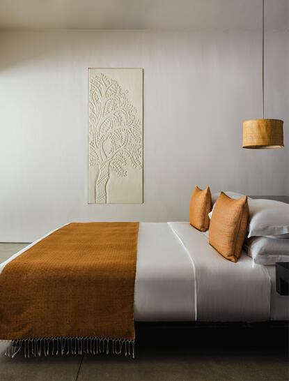 Principal bedroom at Amansara featuring a wooden bed with ochre and cream bedding, wall art, and pendant lighting.