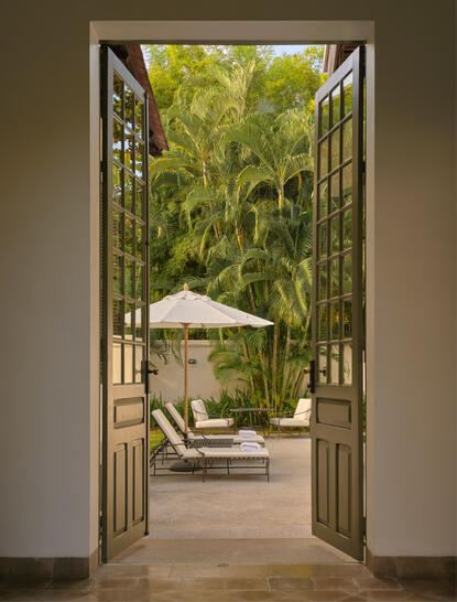 View through open wooden doors to a sunlit courtyard with loungers and parasol at Amantaka.