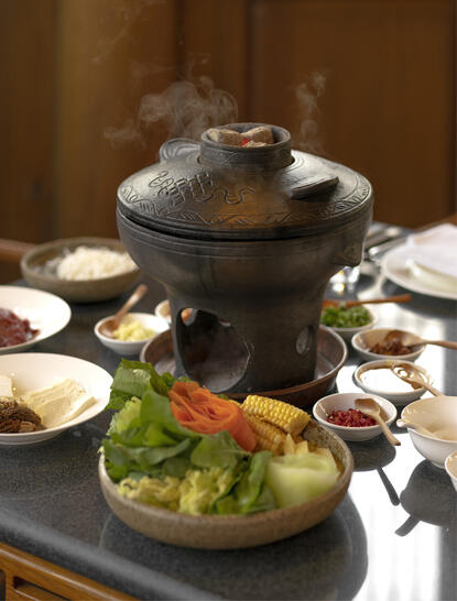 大研安缦度假村的餐饮场景，黑色火锅炉中冒着热气，周围摆放着新鲜食材和陶瓷碗盏。