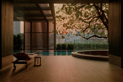 Tranquil swimming pool viewed through floor-to-ceiling windows at Aman Nai Lert Bangkok, with sculptural tree and lounge chair.