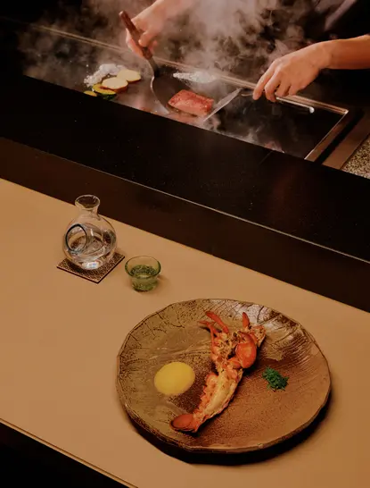 Plated dish with vegetables and garnish at Aman Nai Lert Bangkok, with chef's hands working at the open kitchen counter above.