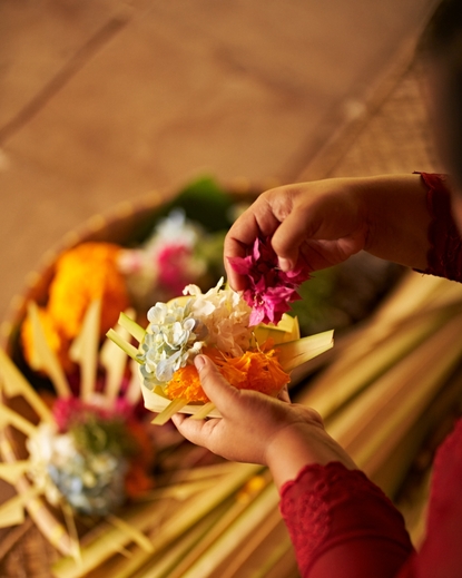 Hands holding a canang sari (Balinese offering) at Amandari, with colourful flowers and palm leaf arrangements.
