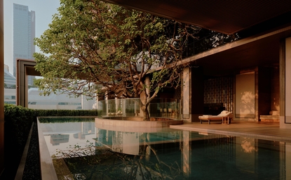 Wellness swimming pool at Aman Nai Lert Bangkok, illuminated at dusk with a large tree and wooden structure nearby.