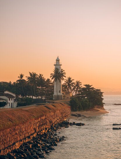 Galle Fort's lighthouse at sunset, with palm trees and rocky shoreline at Amangalla, Sri Lanka.