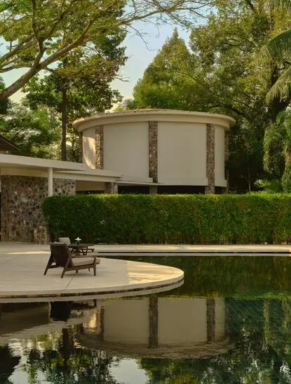 Amansara's main pool and alfresco area reflected in still water, surrounded by verdant landscaping and modern architecture.