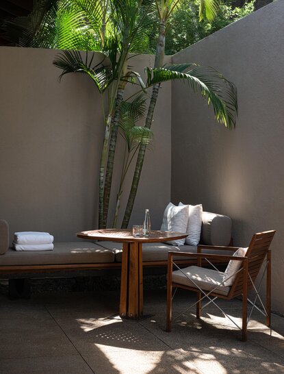 Sunlit corner at Amanwella with potted plant, wooden table, and chair casting shadows on grey wall.